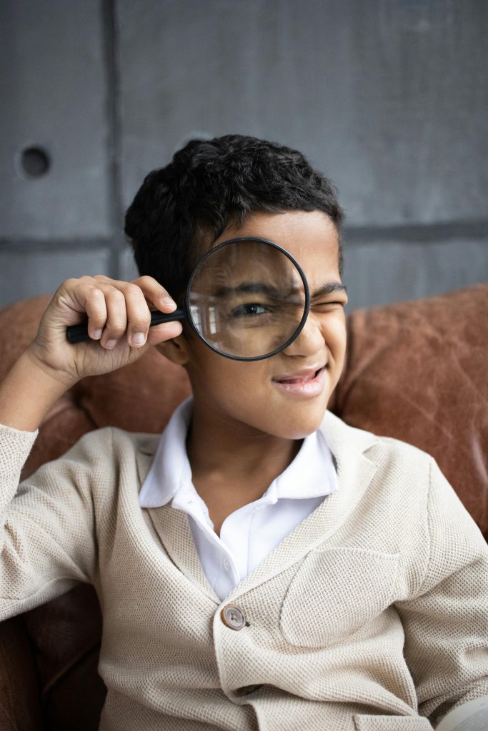 pexels-photo-6186146-6186146 Curious Arabian schoolboy wearing white shirt with cardigan sitting on comfortable couch and looking through magnifying glass at camera indoors